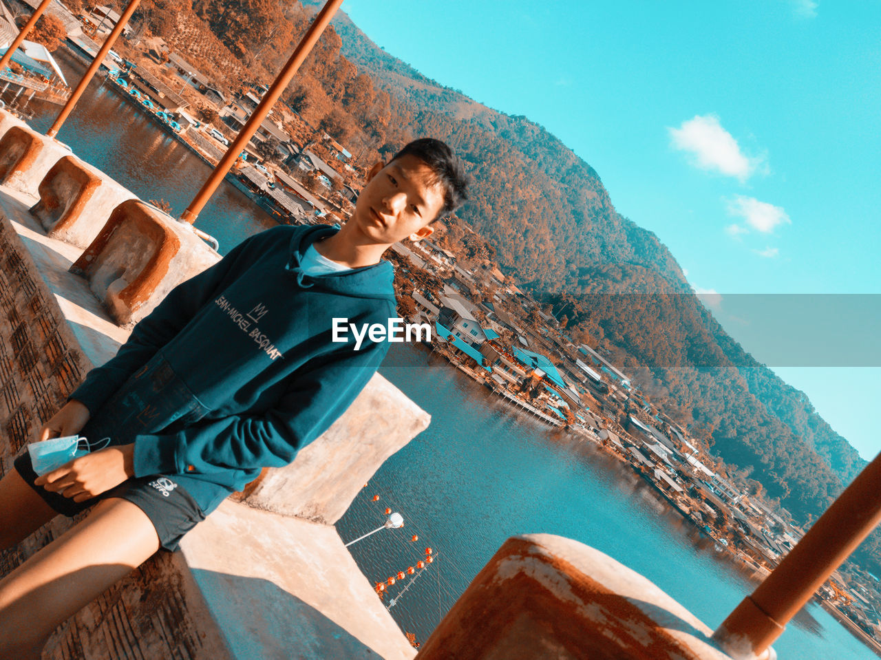 YOUNG MAN SITTING ON SHORE AGAINST MOUNTAINS AGAINST SKY