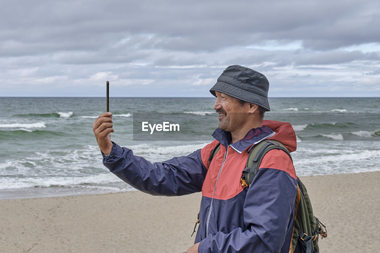 Man standing at beach and shooting video on phone against sea and sky