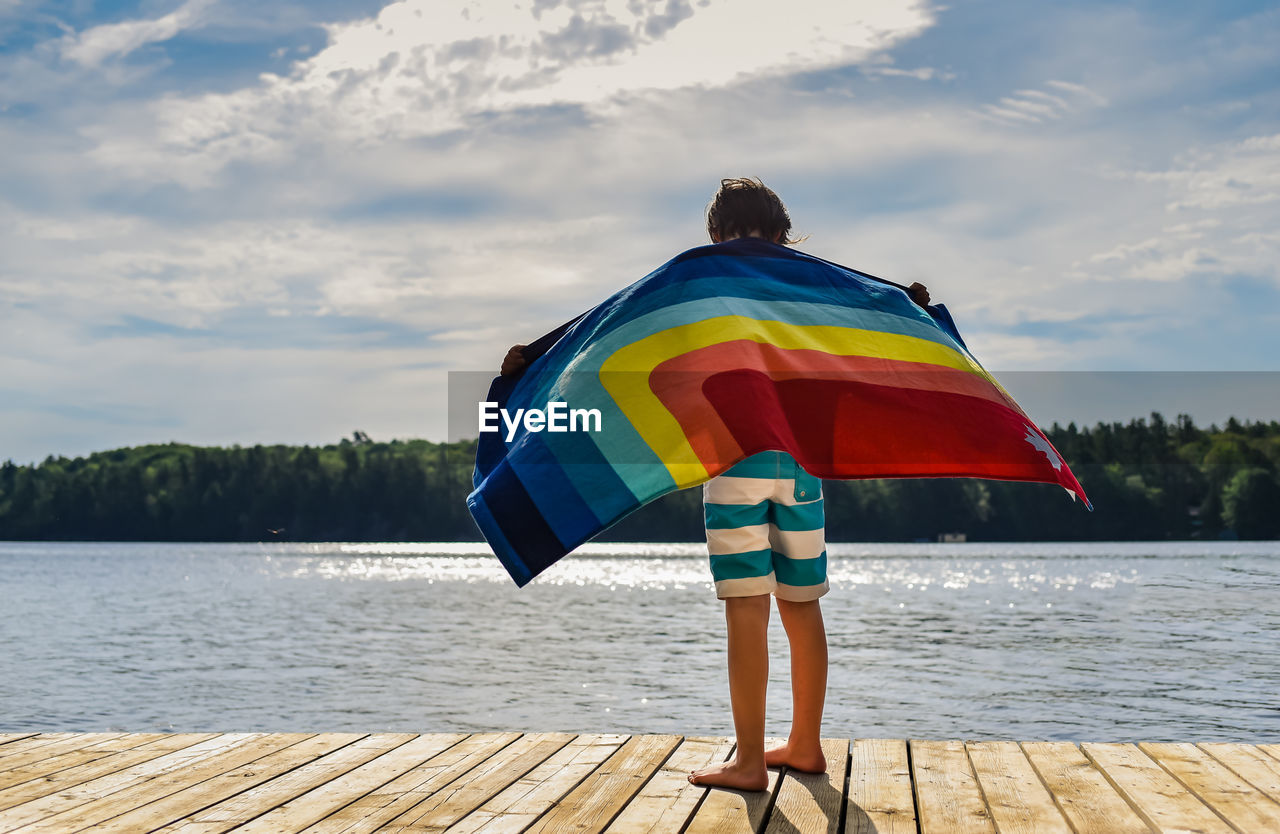 Child standing on a dock holding a rainbow towel in the wind.