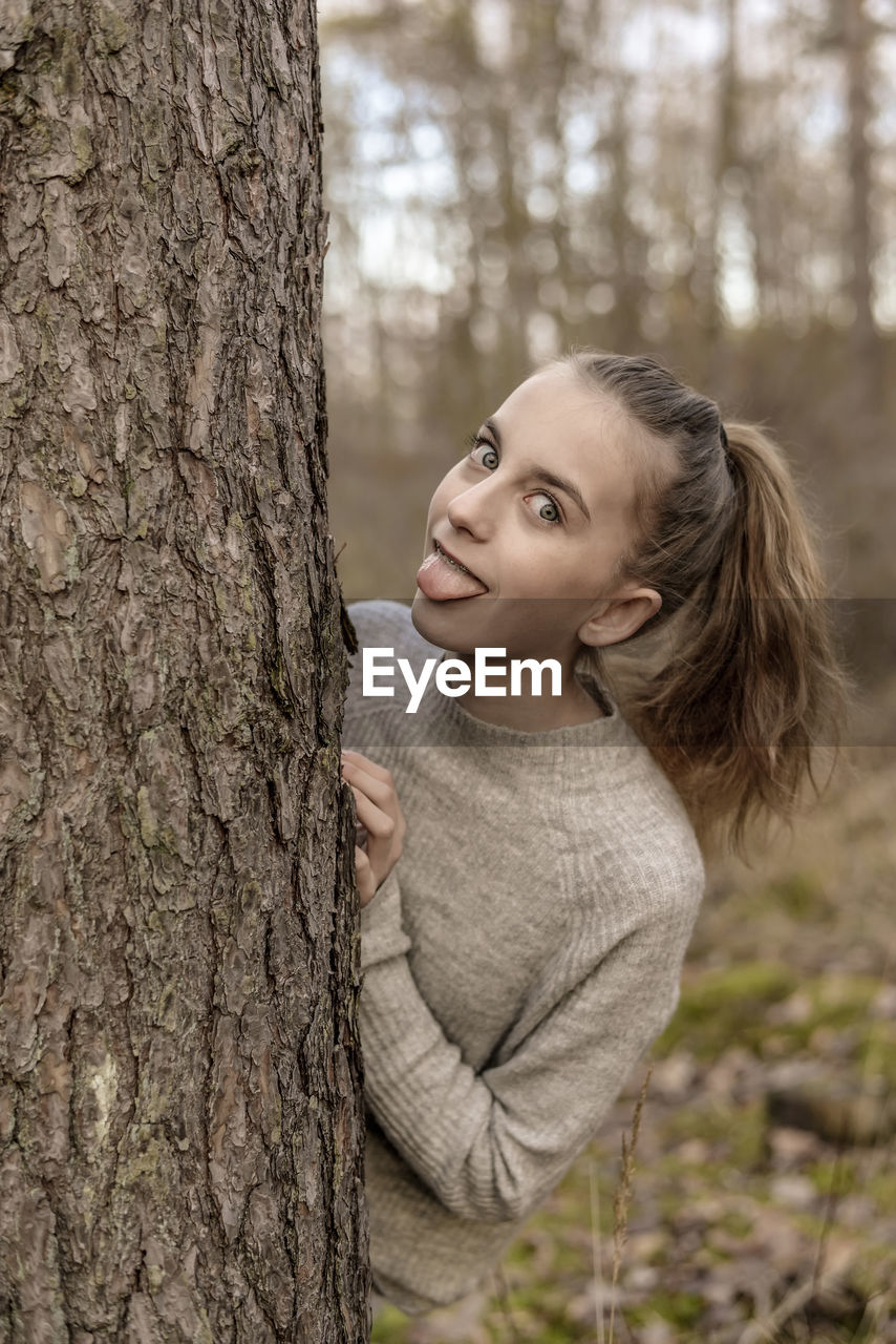 Portrait of teenage girl sticking out tongue while standing behind tree in forest