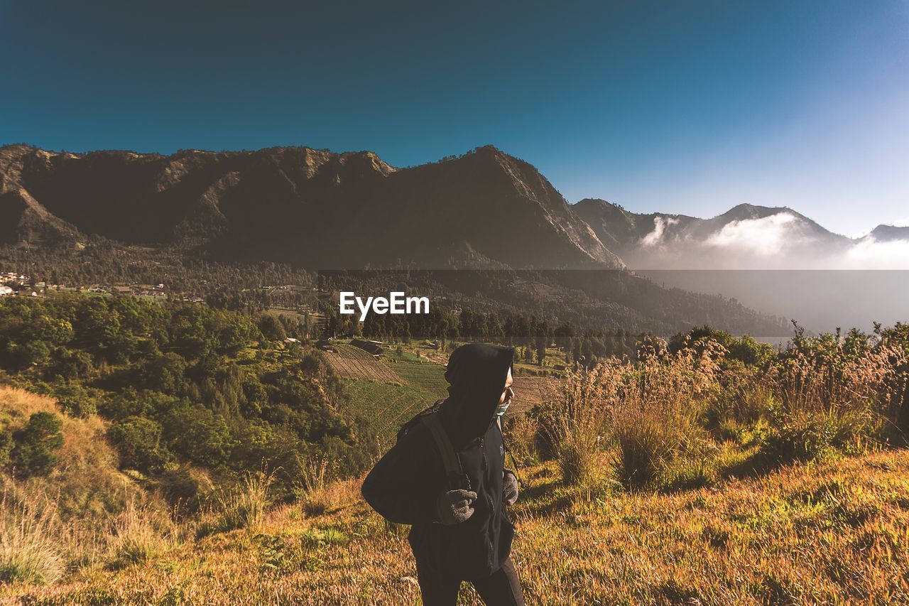 MAN STANDING BY PLANTS AGAINST MOUNTAIN RANGE