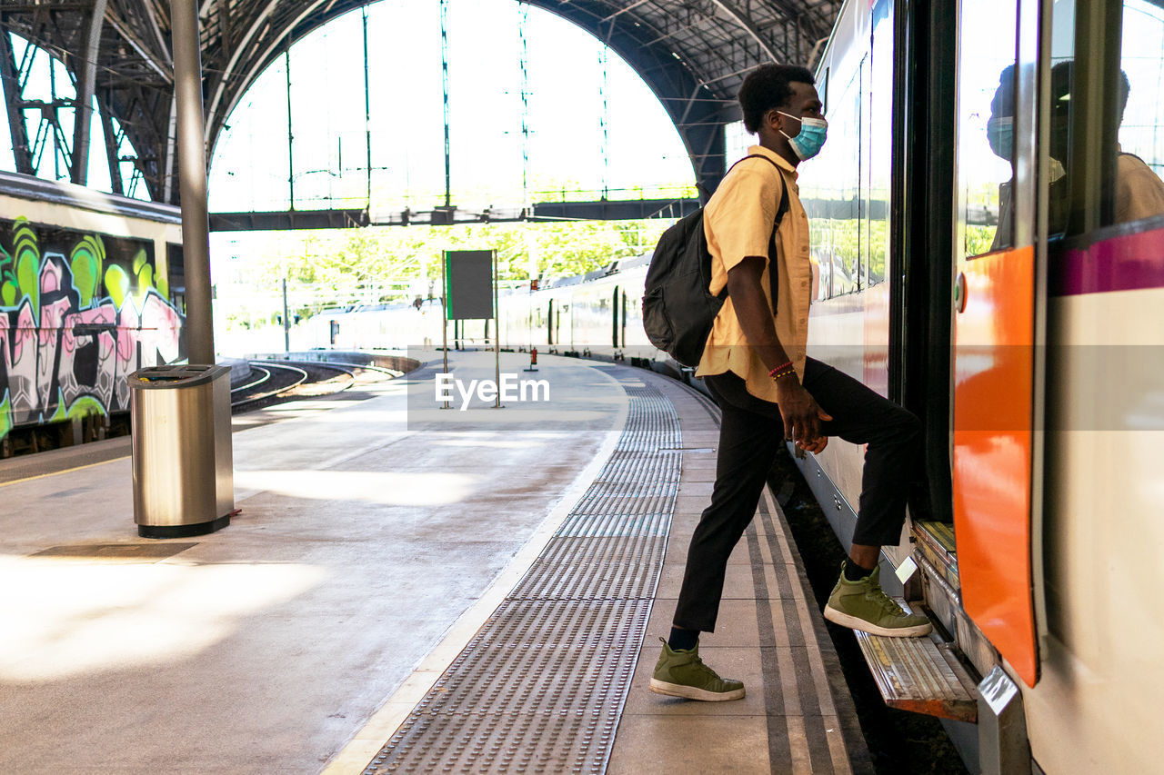 Side view of african american male wearing medical mask entering modern train on station during coronavirus epidemic