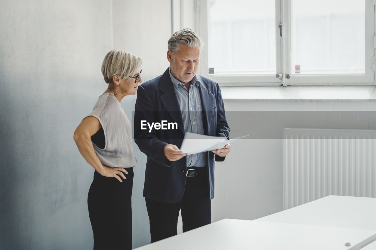 Mature businessman reading documents while discussing with female colleague in new office