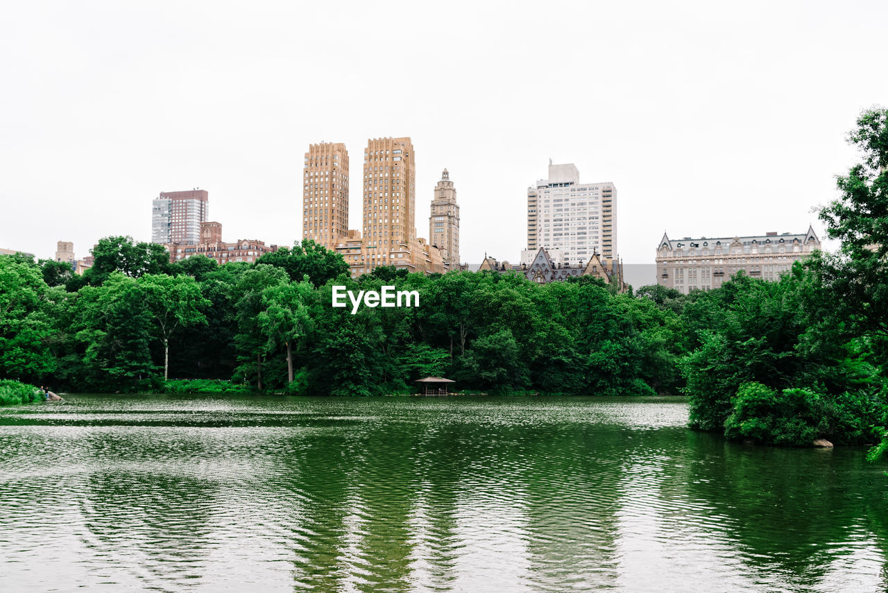 Trees by lake against buildings in city against sky