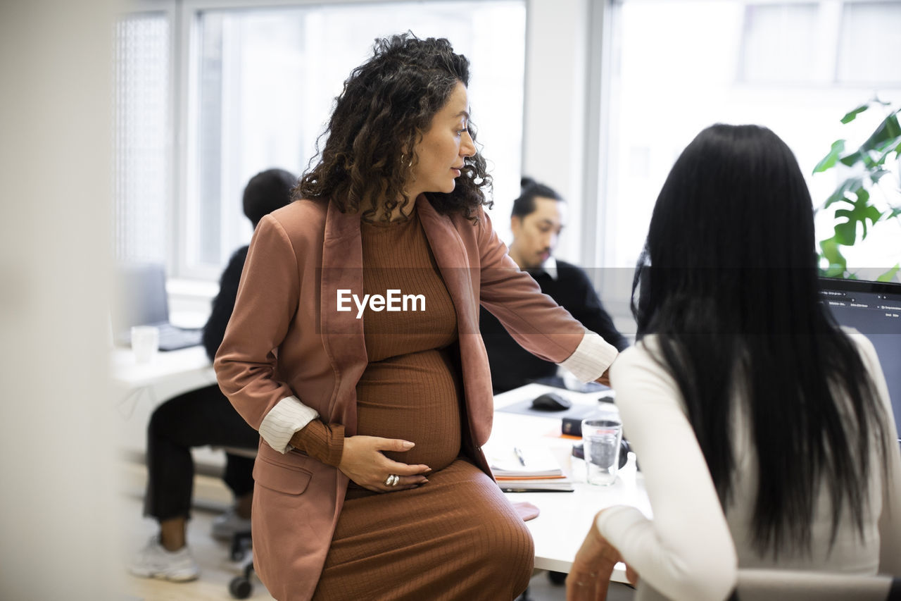 Pregnant businesswoman discussing with female colleague over computer in office