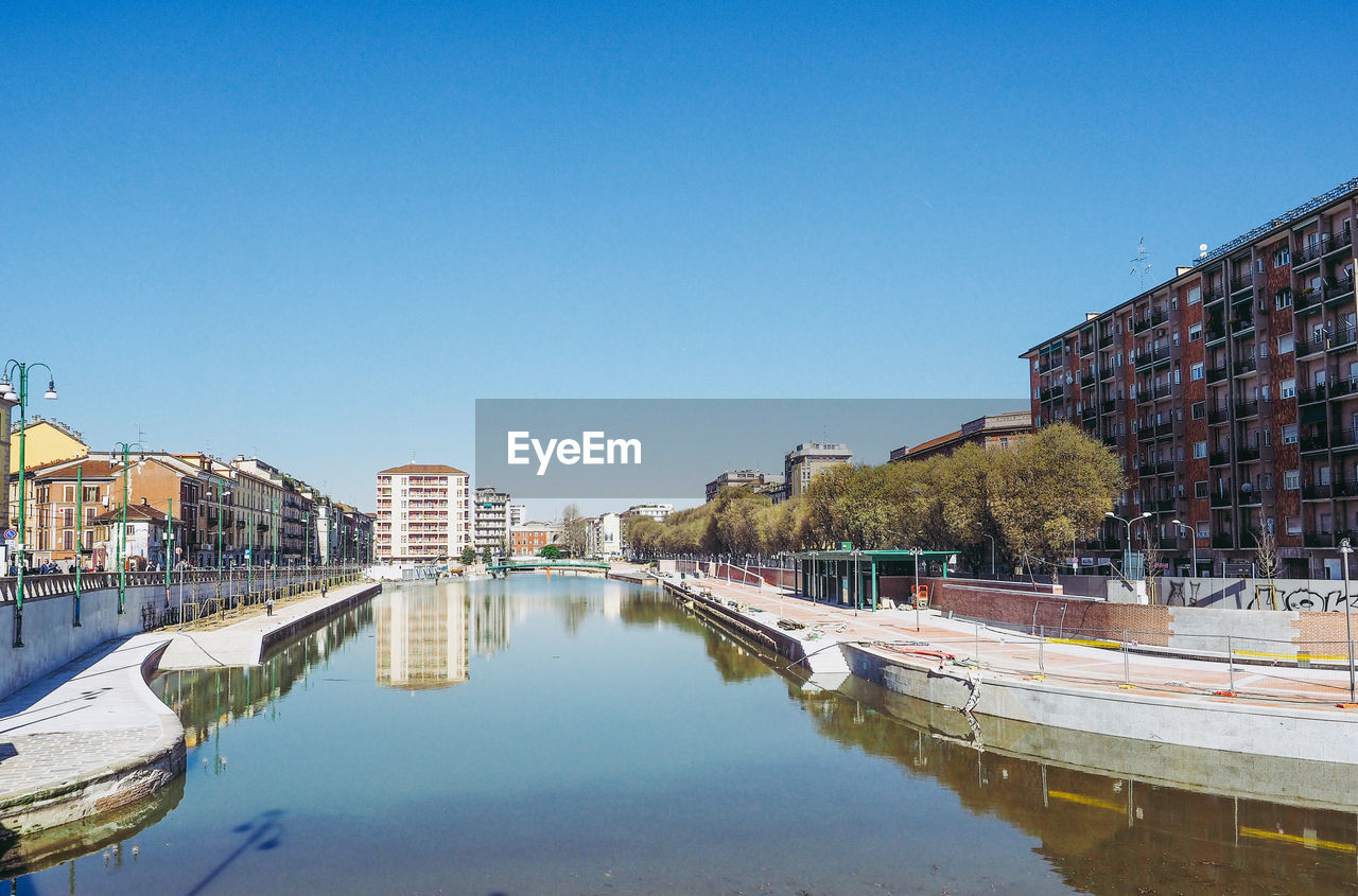 Reflection of buildings and trees in canal against clear blue sky