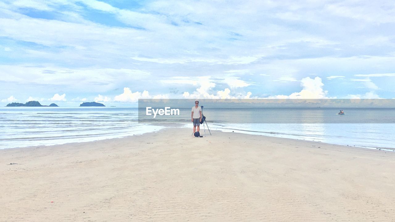 Man standing with tripod at beach against cloudy sky