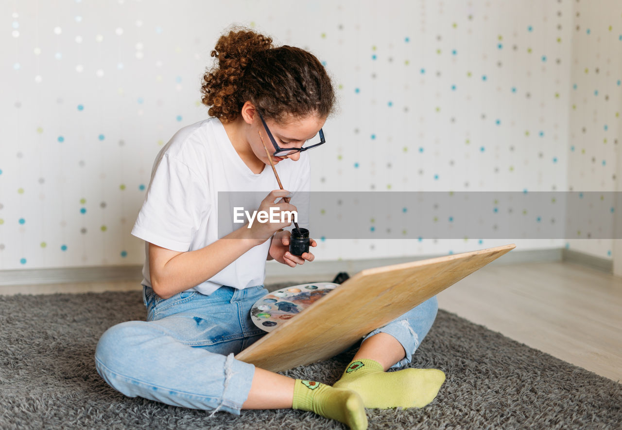 Curly hair girl teenager in glasses in white t-shirt sitting on floor painting on wooden desk 