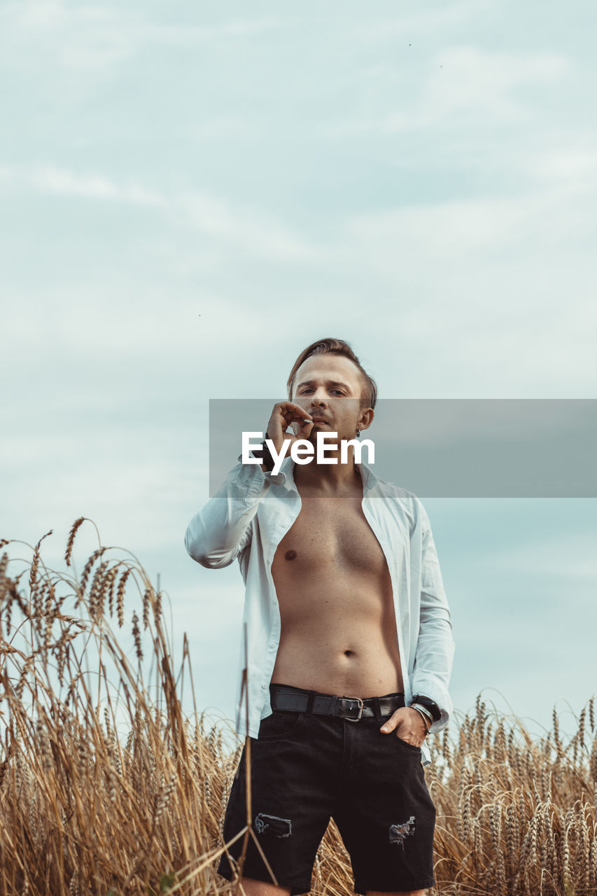 Young man smoking while standing on field against sky