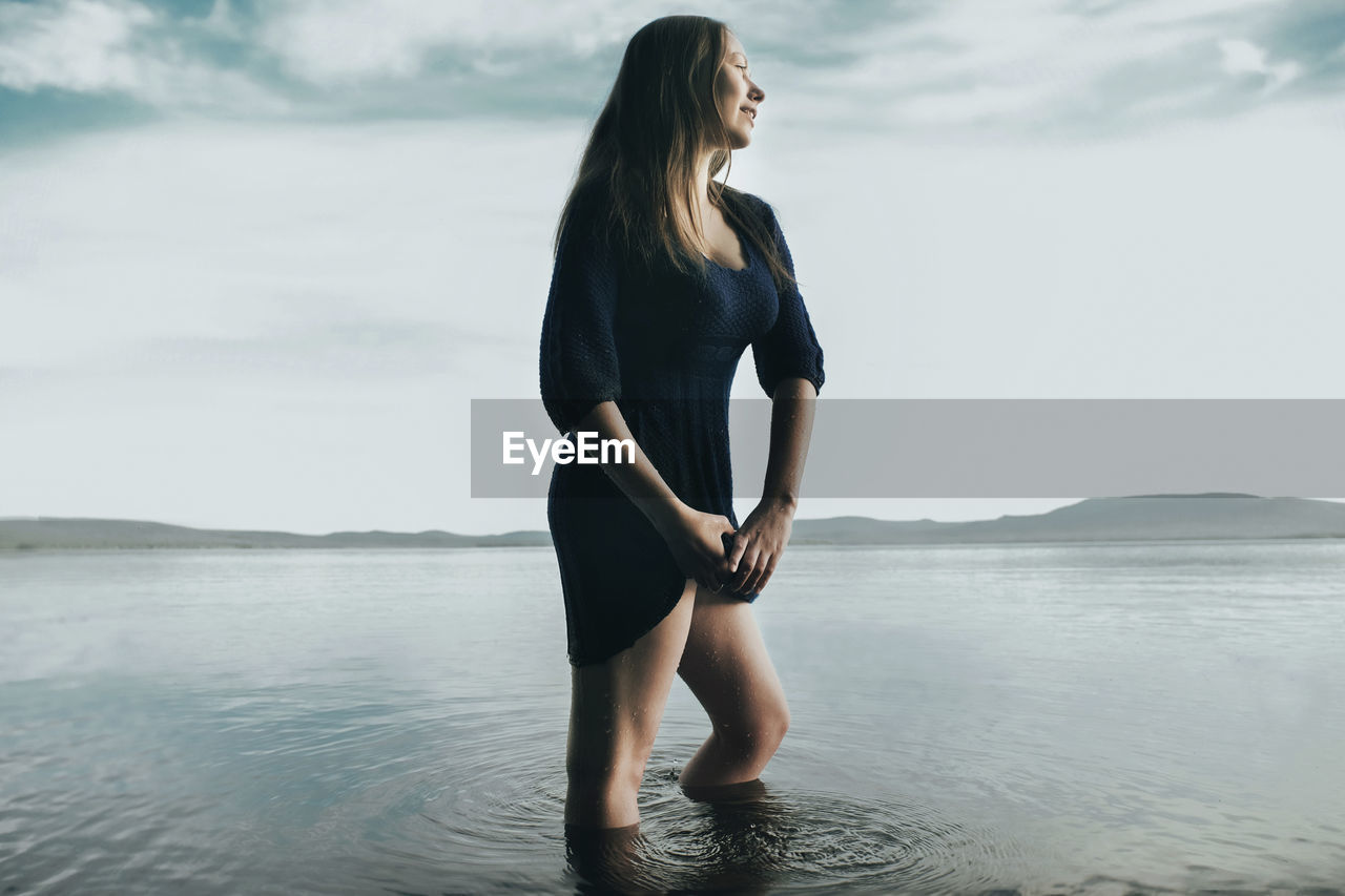 Thoughtful woman squeezing dress while standing in sea against sky