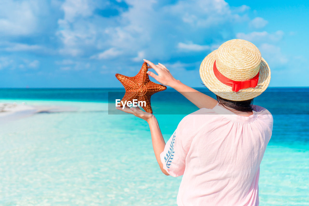 Rear view of woman holding starfish on beach against sky