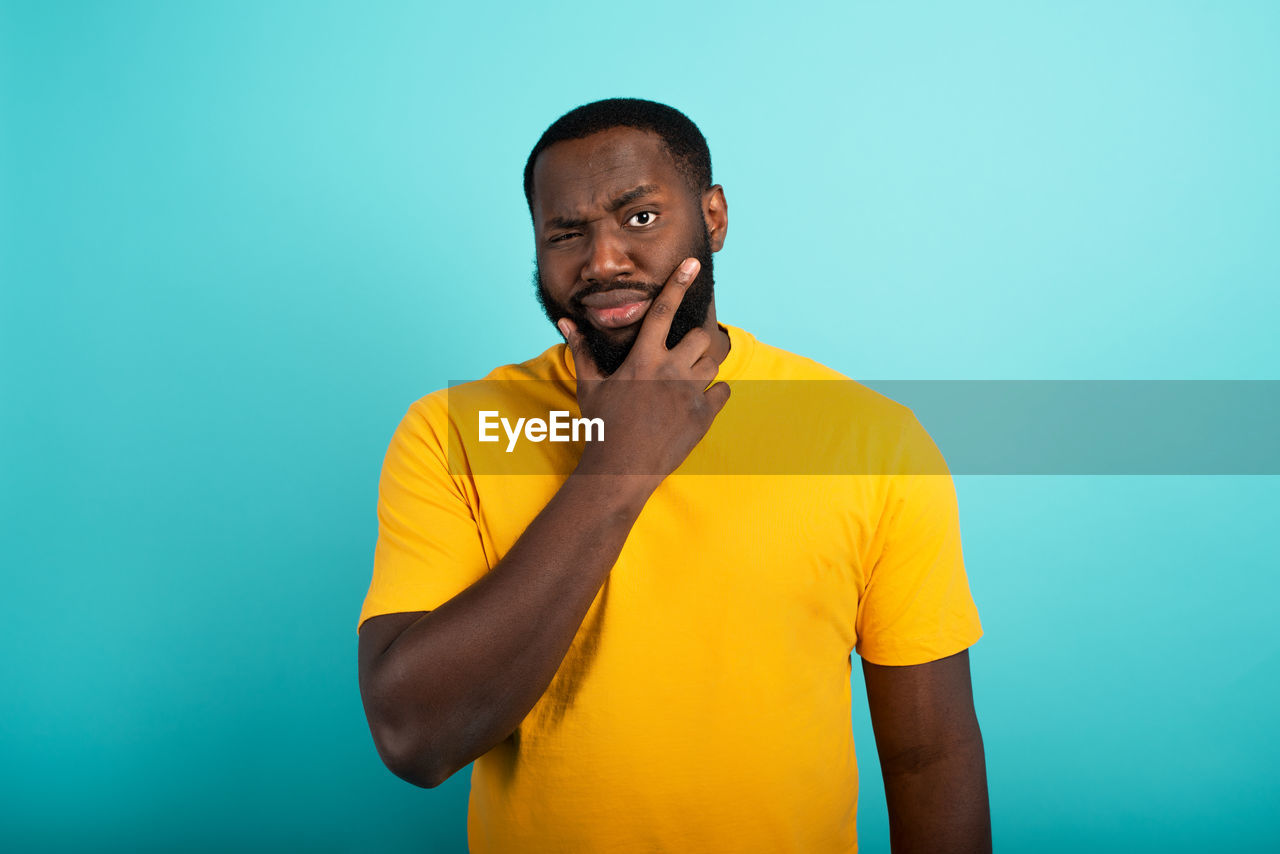 yellow, one person, colored background, adult, studio shot, men, portrait, waist up, emotion, indoors, shaved head, casual clothing, copy space, standing, person, happiness, smiling, t-shirt, young adult, blue, lifestyles, looking, front view, finger, looking at camera, blue background, facial hair, beard