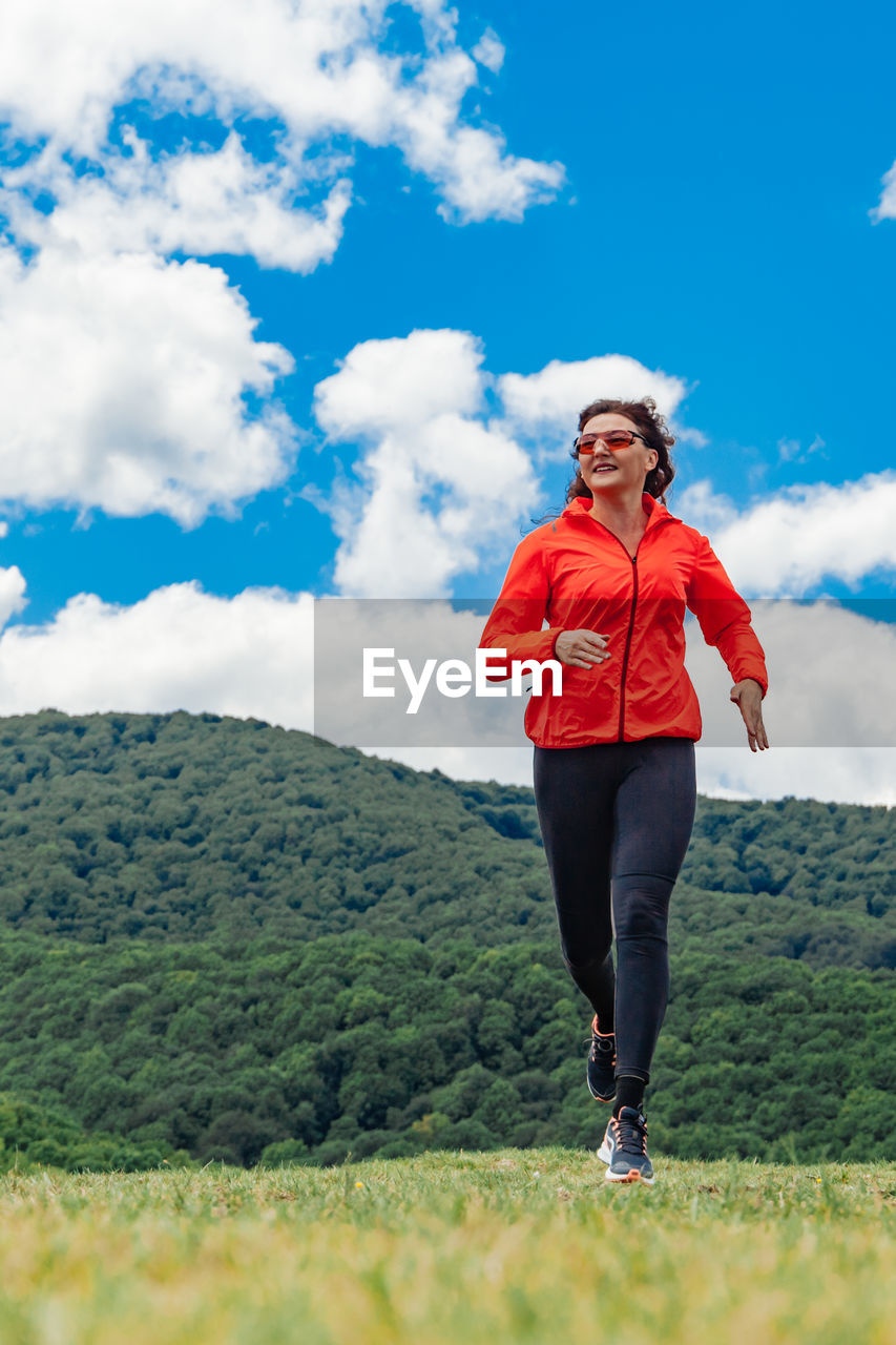 Adult woman in a bright sports jacket and leggings does a morning jog on green hill against a sky