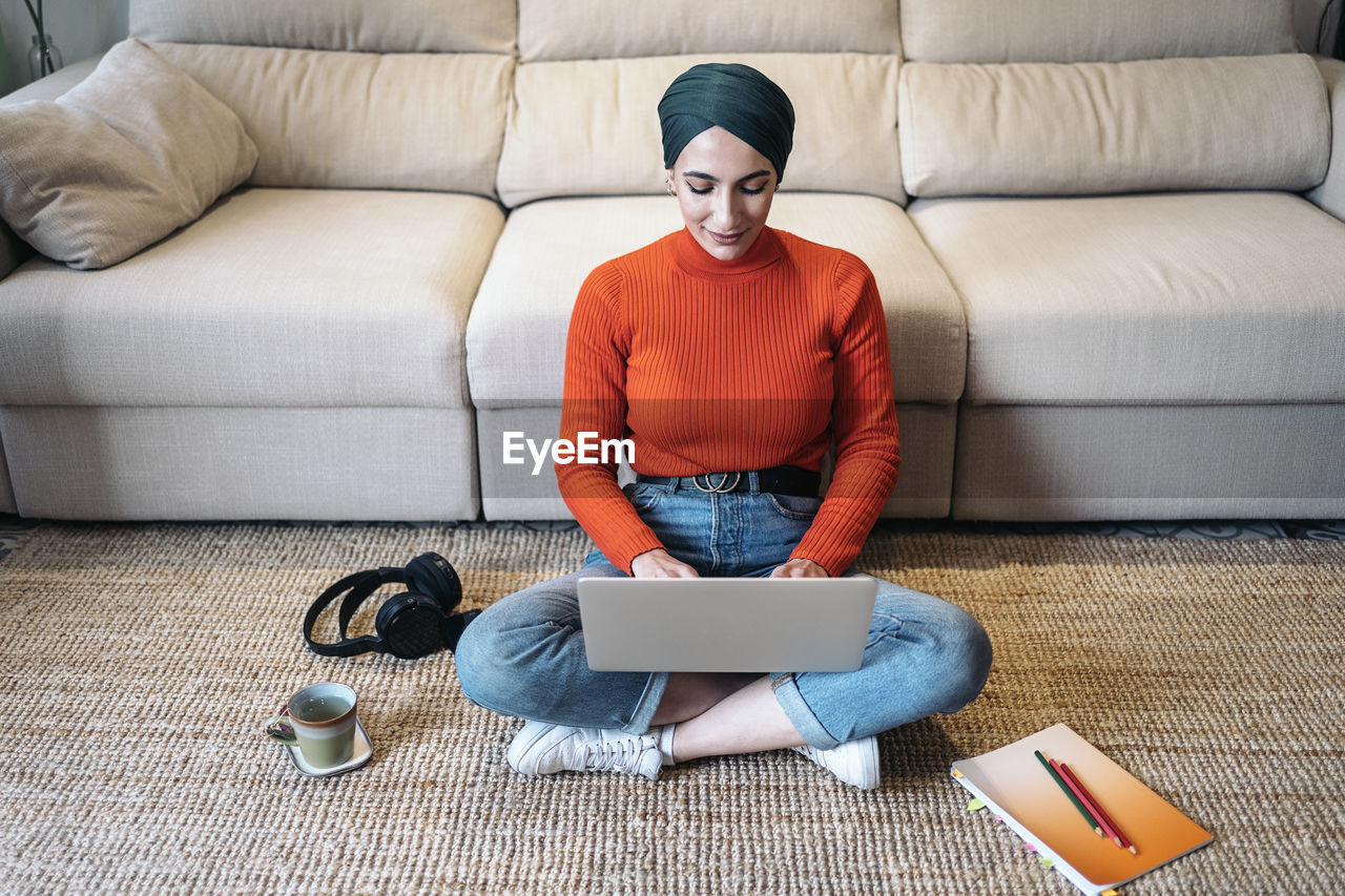 High angle of glad muslim woman in casual clothes sitting cross legged on floor near sofa and browsing data on netbook while working on remote project at home