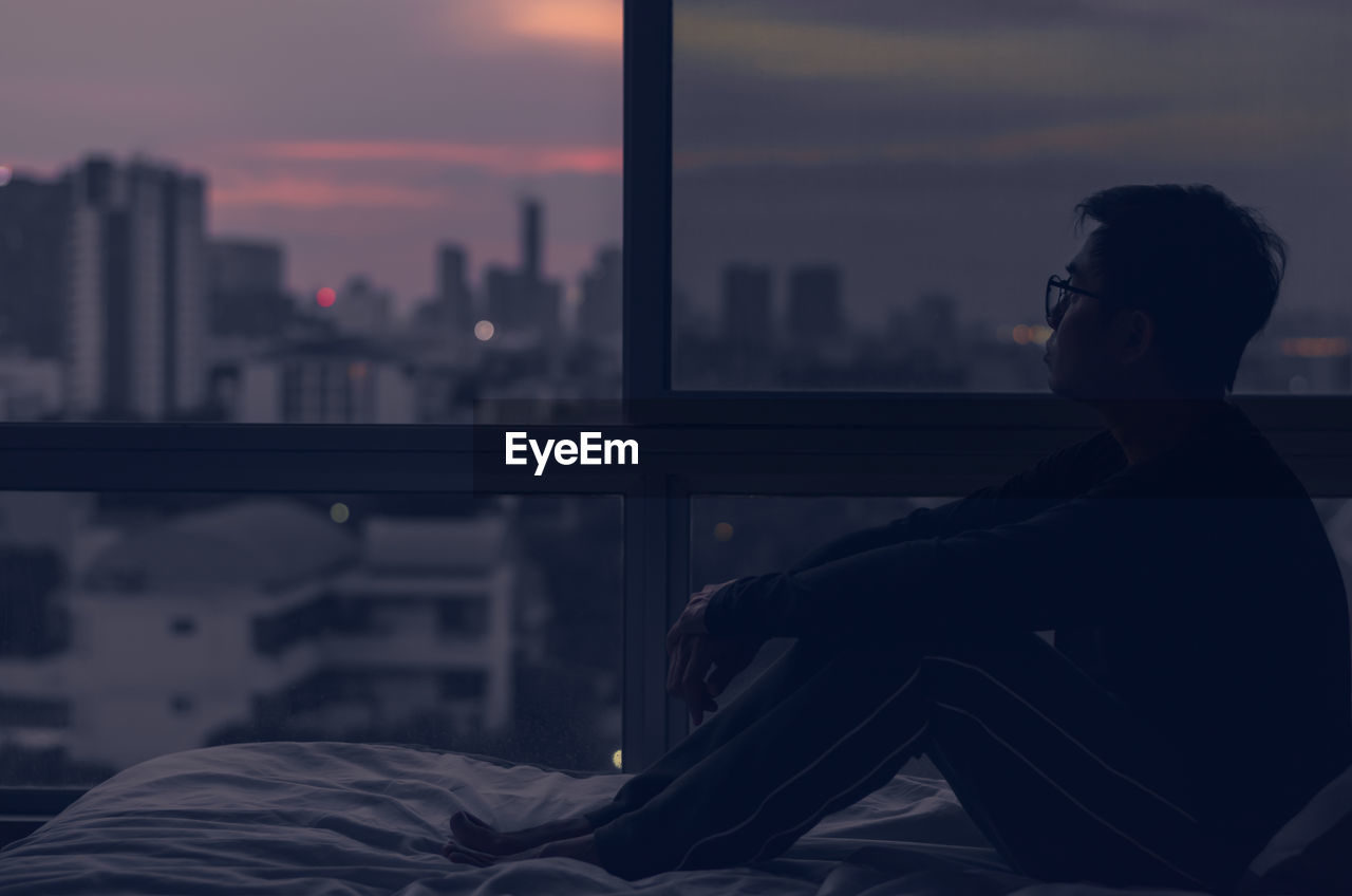 The man sitting on bed with city view from bedroom. stay home, depression and loneliness concept.