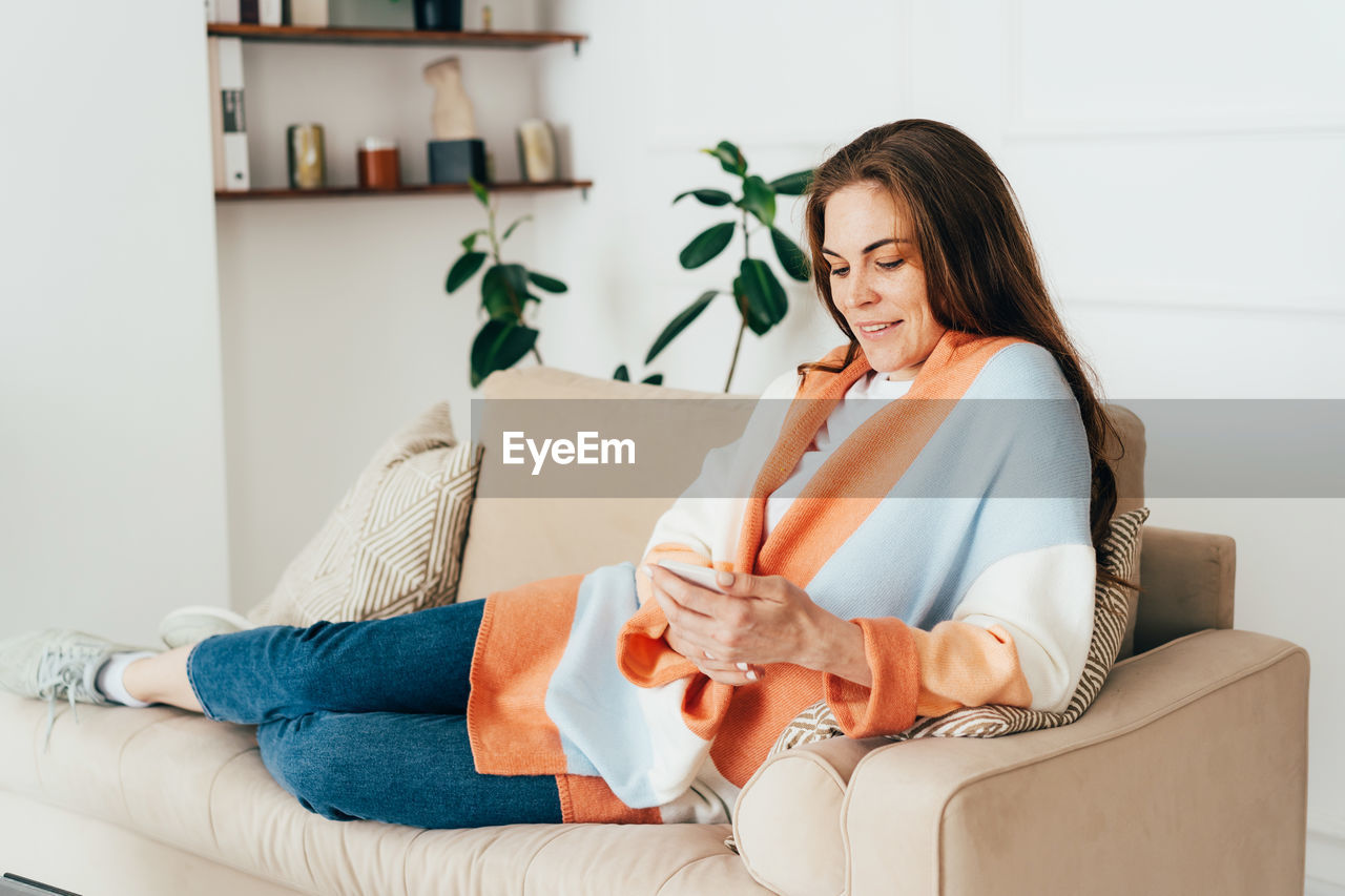 Young redhead woman sitting on the sofa in the living room uses the phone to communicate.