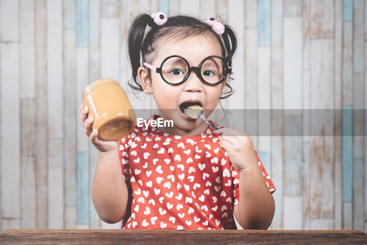 Portrait of girl eating peanut butter at table against wall