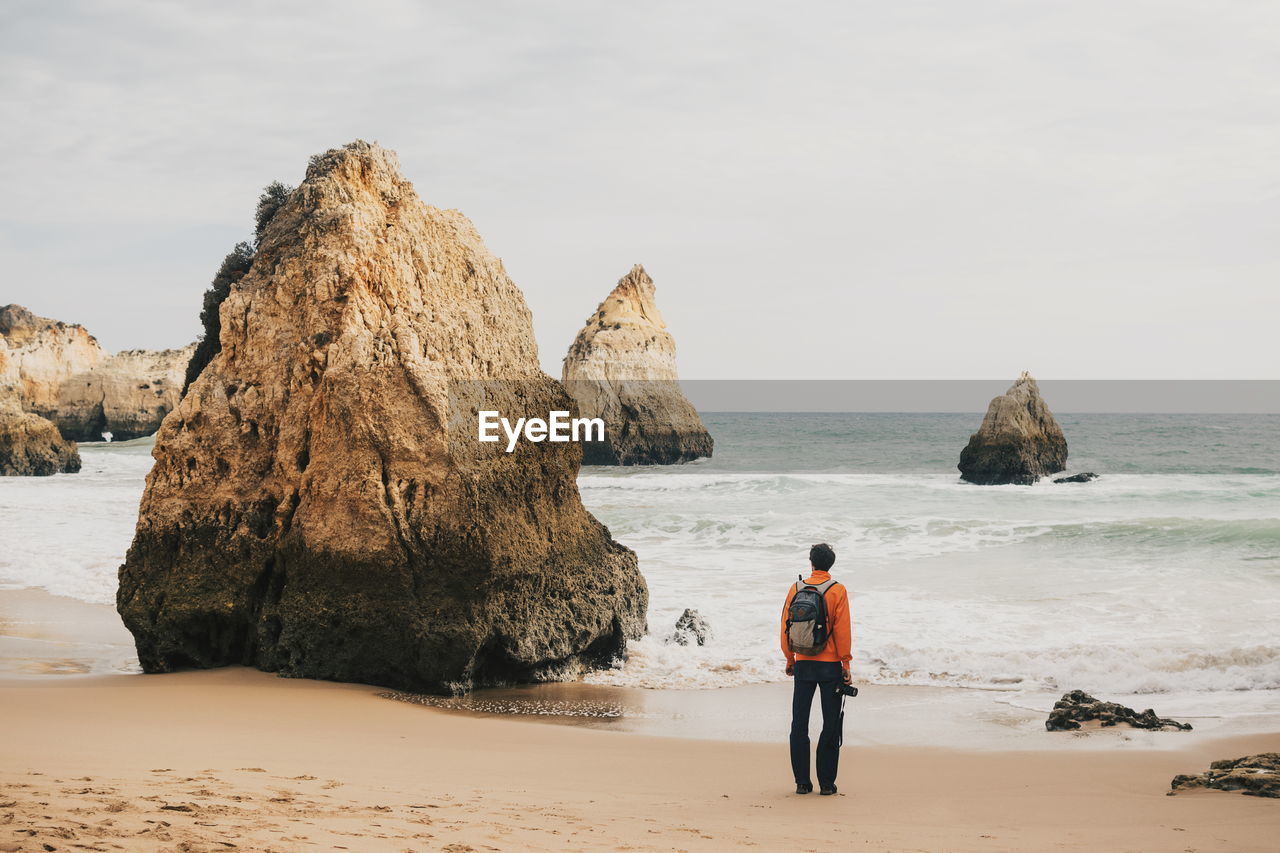 Rear view of man standing by rock at beach against sky