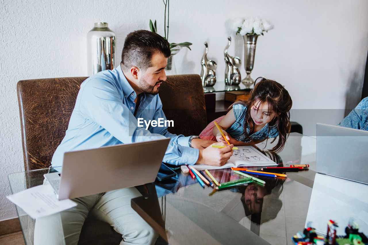 Young latin american self employed man helping cute little daughter to paint in album with colorful pencils while sitting at table and working remotely on laptop