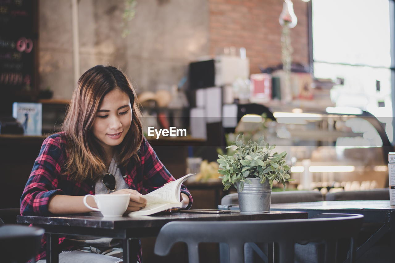 Smiling young woman reading book while sitting by coffee on table at cafe