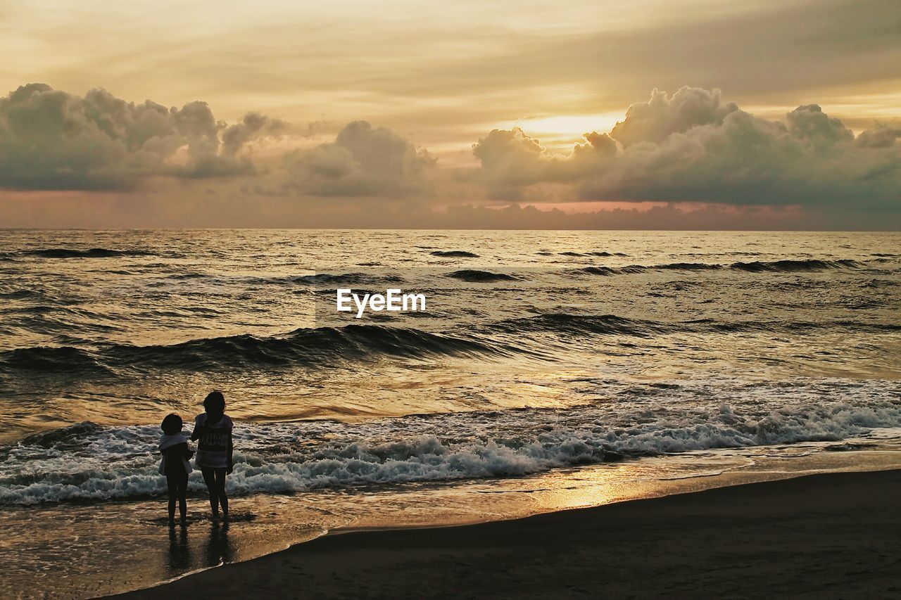 Girls standing at shore against sky during sunset