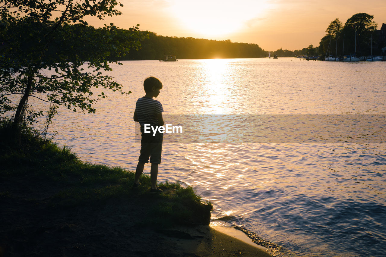 Rear view of boy standing by lake during sunset