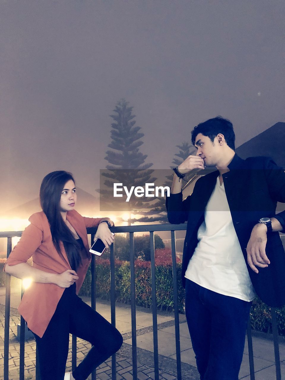 Couple looking at each other while standing by railing against sky at night