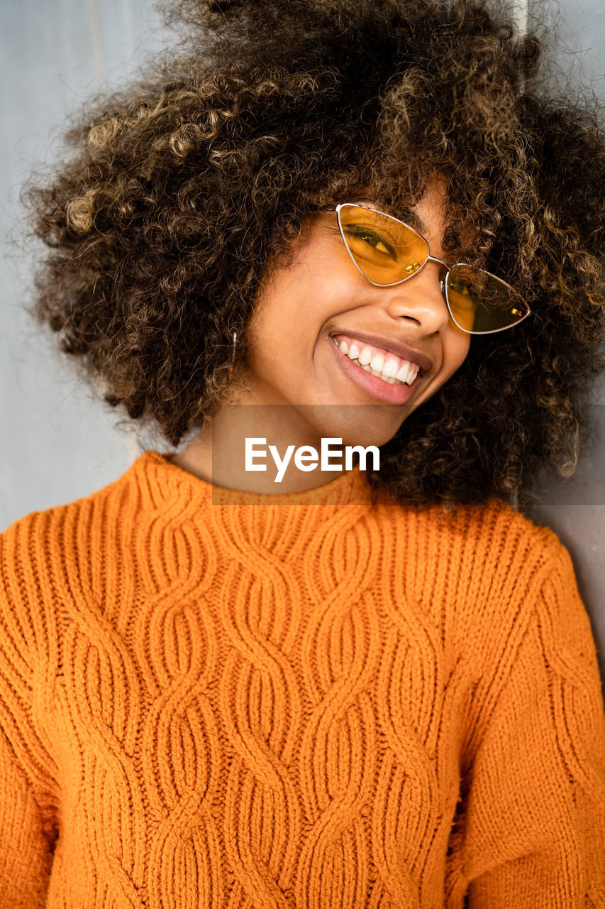 Beautiful cheerful ethnic woman with curly afro hair wearing vivid orange sweater looking at camera against gray wall