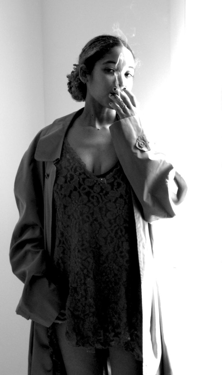 Portrait of young woman wearing trench coat smoking while standing against wall