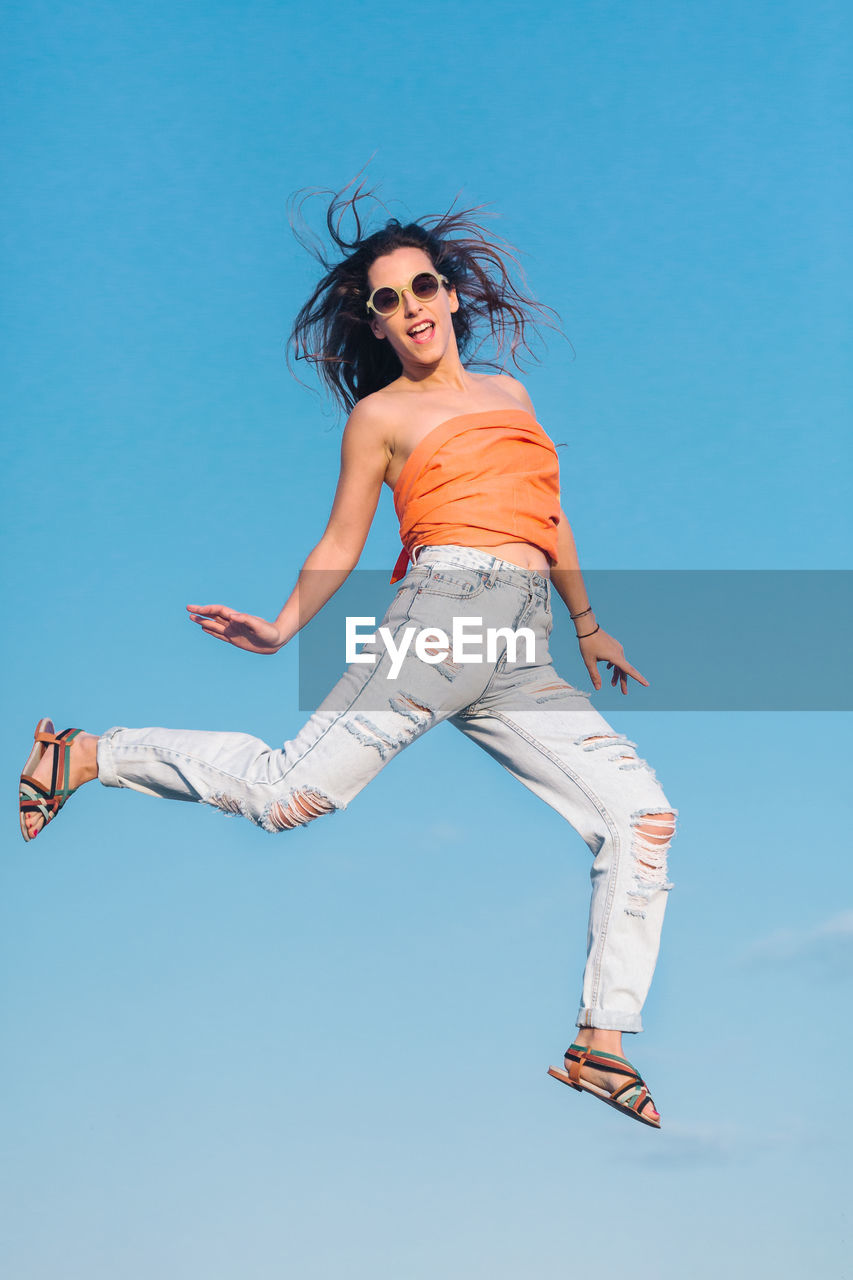 From below full body of modern hipster female in sunglasses wearing shirt and stylish ripped jeans jumping high against blue sky