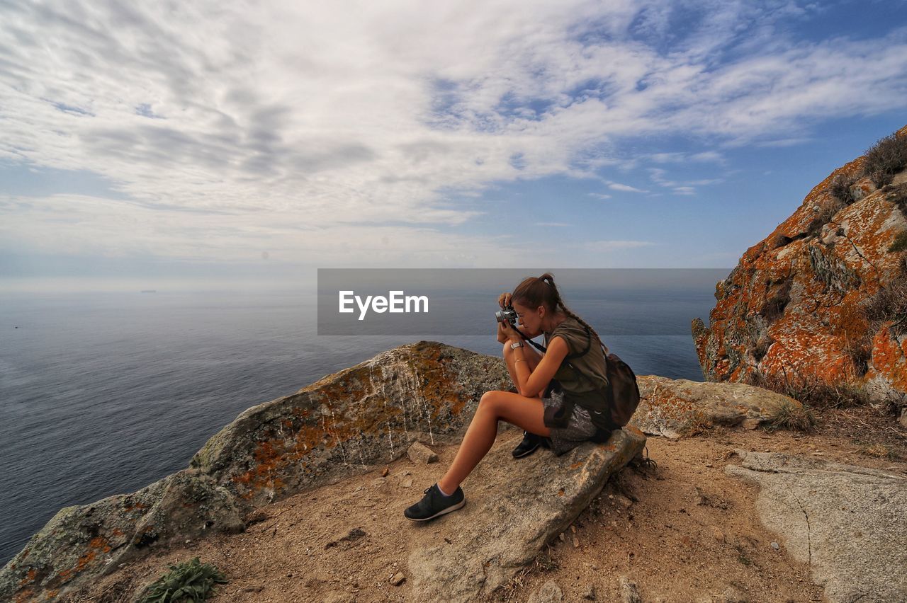 YOUNG WOMAN SITTING ON ROCK LOOKING AT SEA