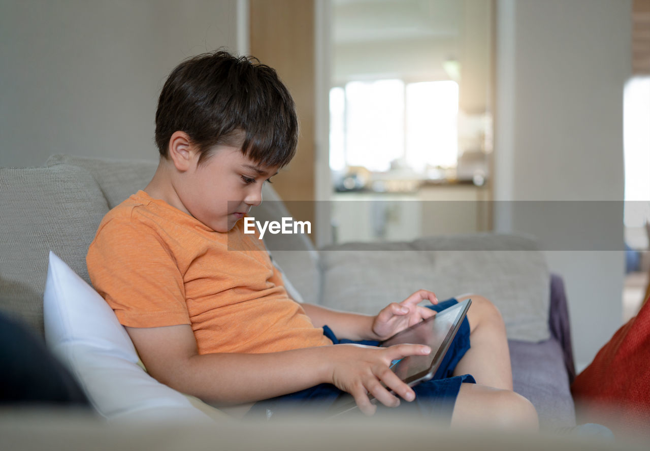 Kid sitting on sofa using tablet learning online on internet, child boy playing game on touch pad,