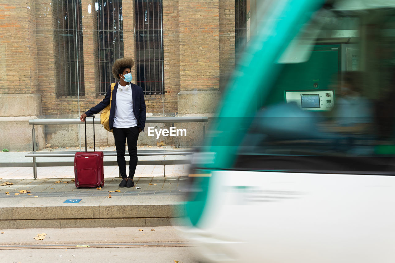 Black man with afro hair and face protective mask standing on the street with a suitcase and a bag hanging from his shoulder in front of a tram that is moving