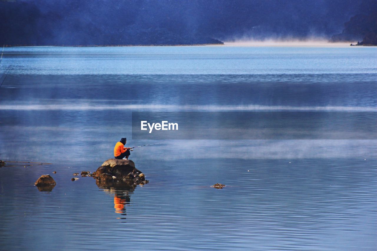 Man fishing in lake while sitting on rock