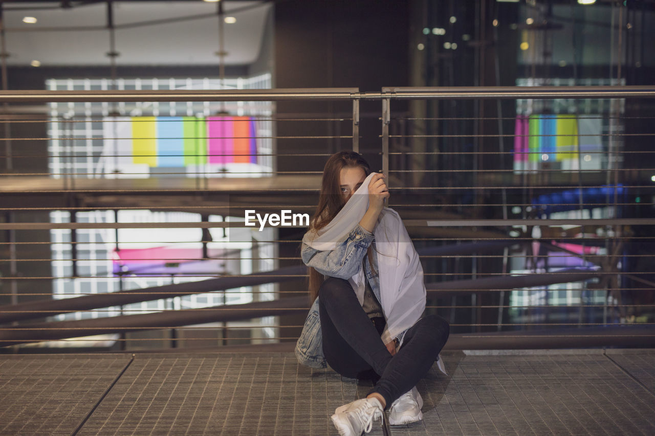 Woman holding fabric over face while sitting on footbridge against railing