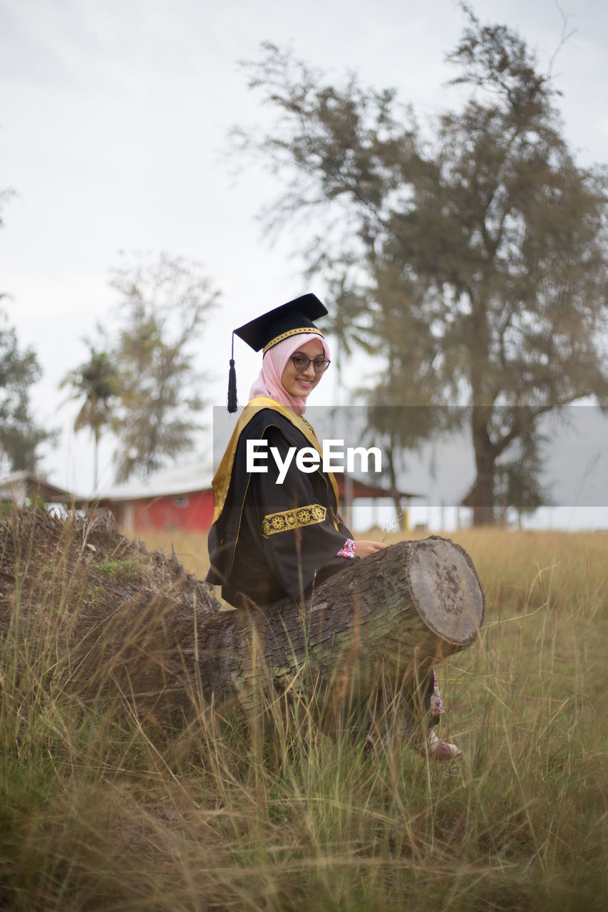 Portrait of woman in graduation gown sitting on log