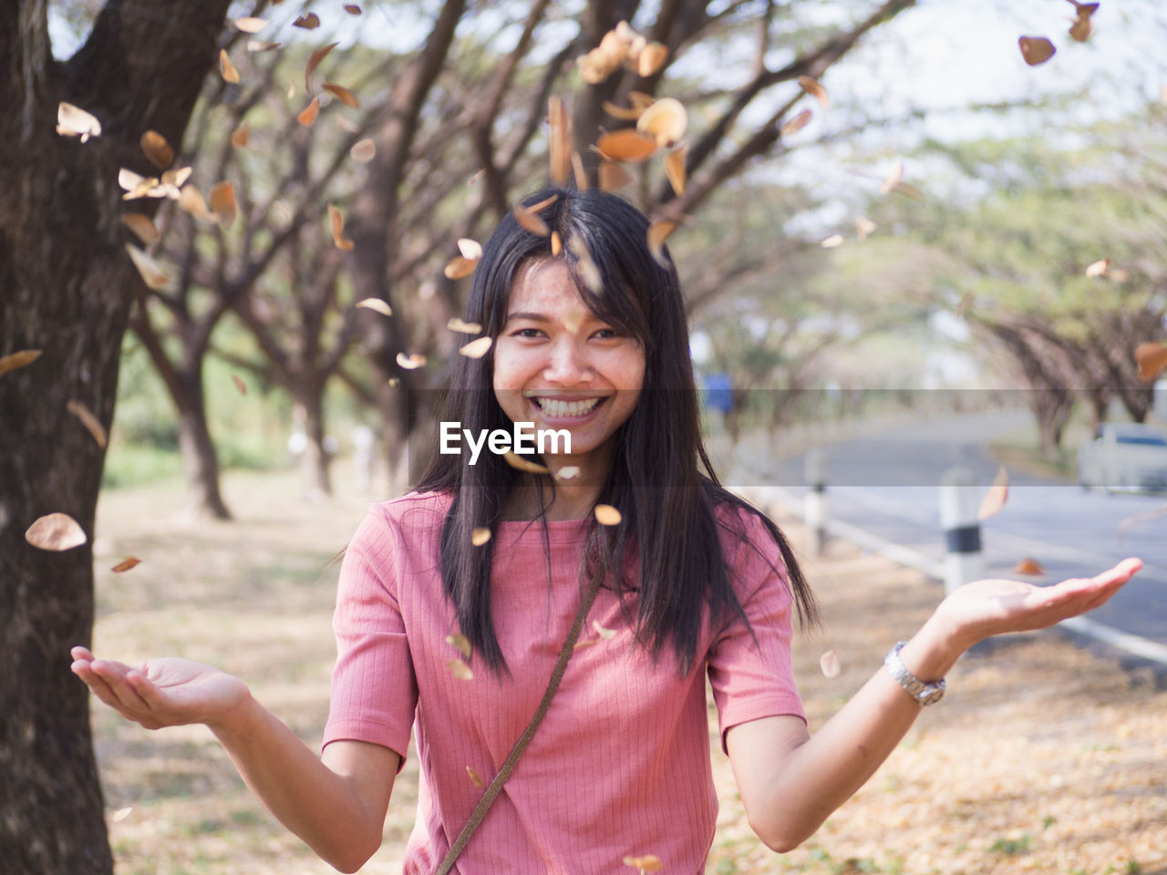 Portrait of smiling young woman throwing leaves and twigs while standing against trees