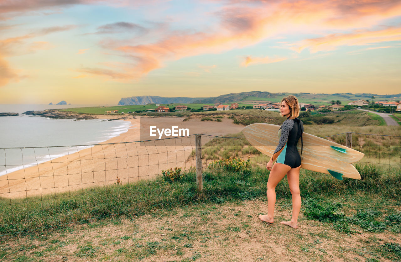 Surfer woman with wetsuit and surfboard looking at the beach