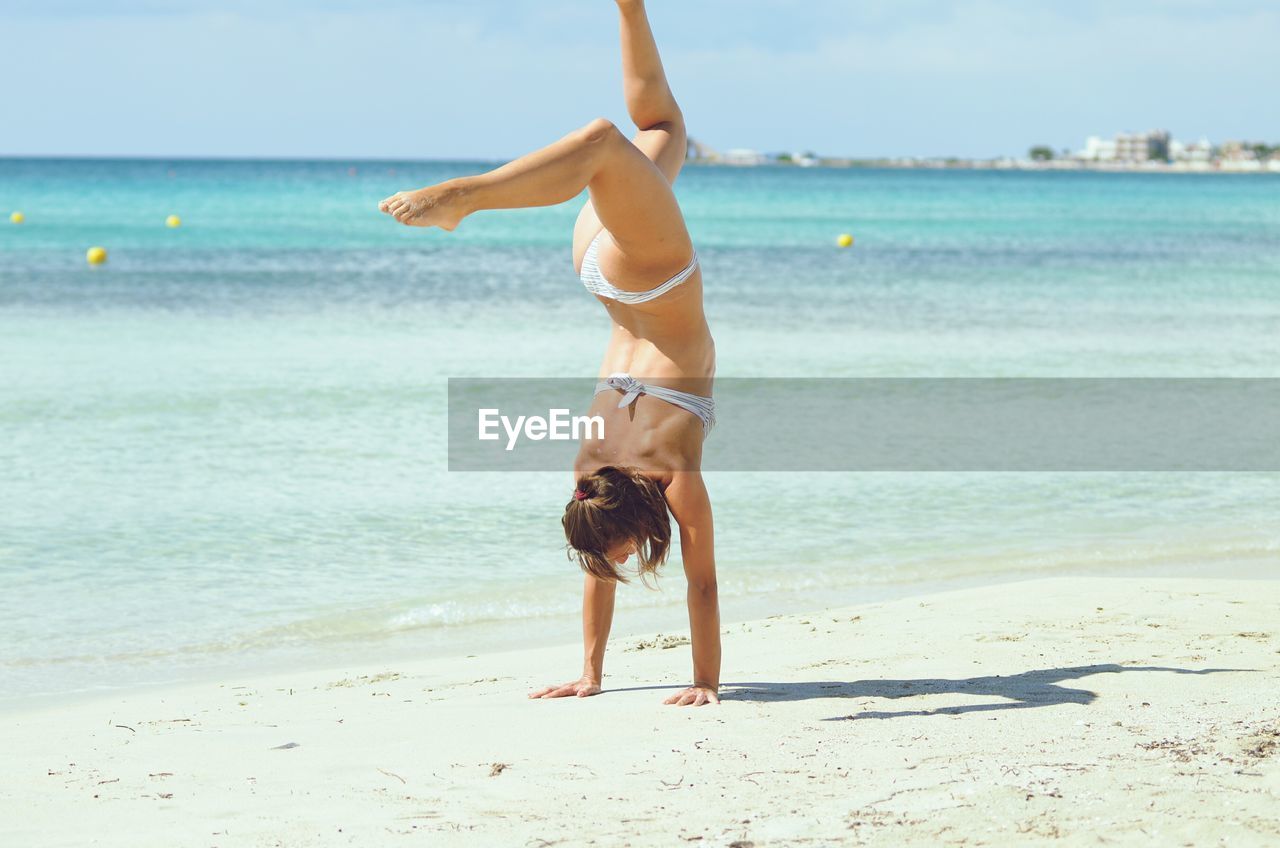 Young woman wearing bikini while practicing handstand at beach during sunny day