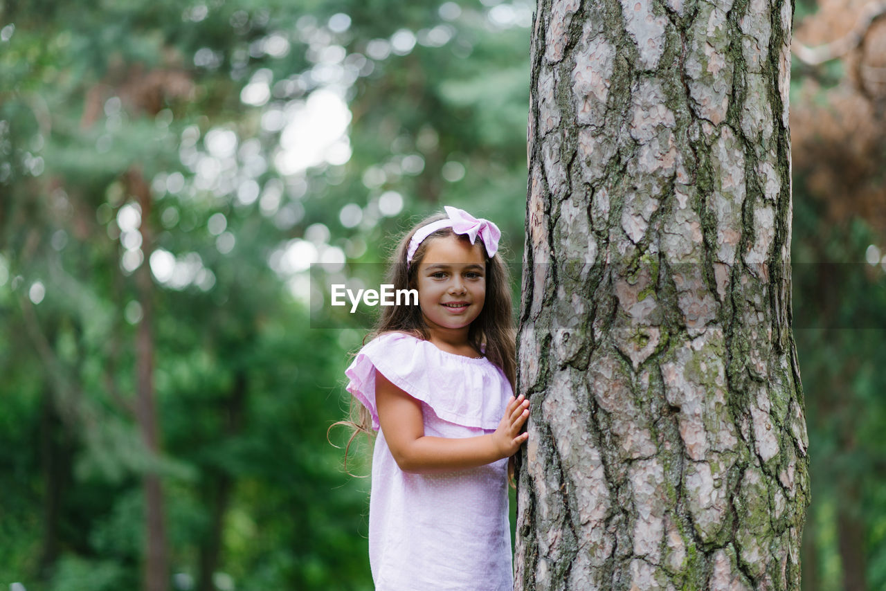 A beautiful girl in a pink dress stands near a tree in the forest in summer, smiling and dreaming