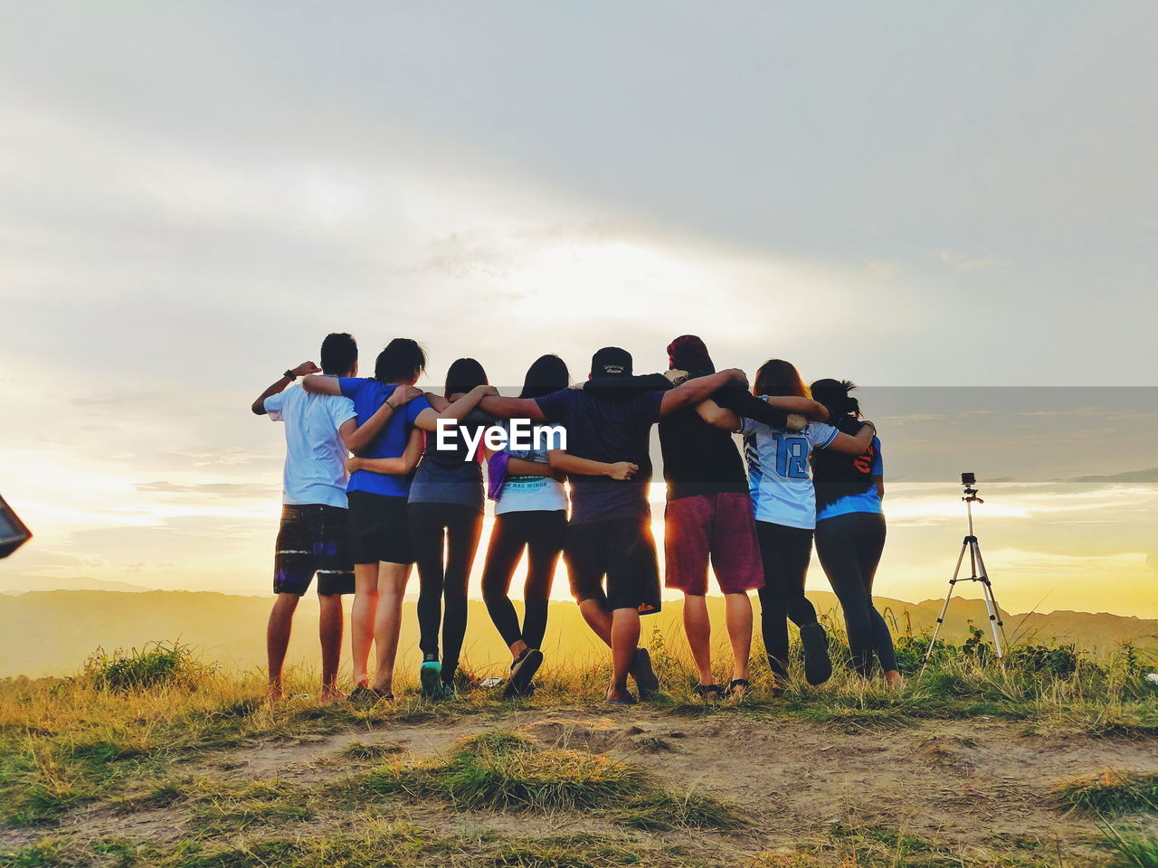 REAR VIEW OF PEOPLE STANDING ON GRASS AGAINST SKY DURING SUNSET