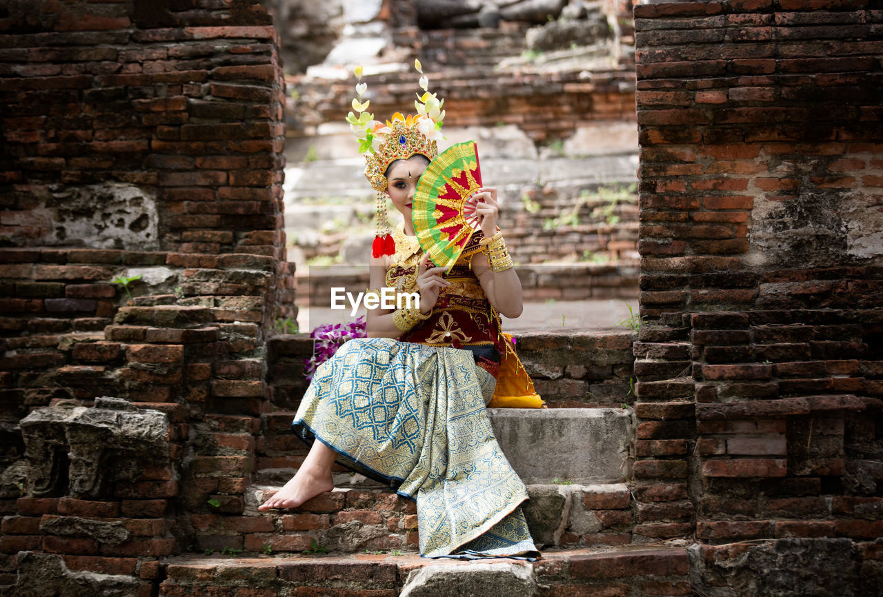 Young woman in traditional clothing holding hand fan while sitting at temple