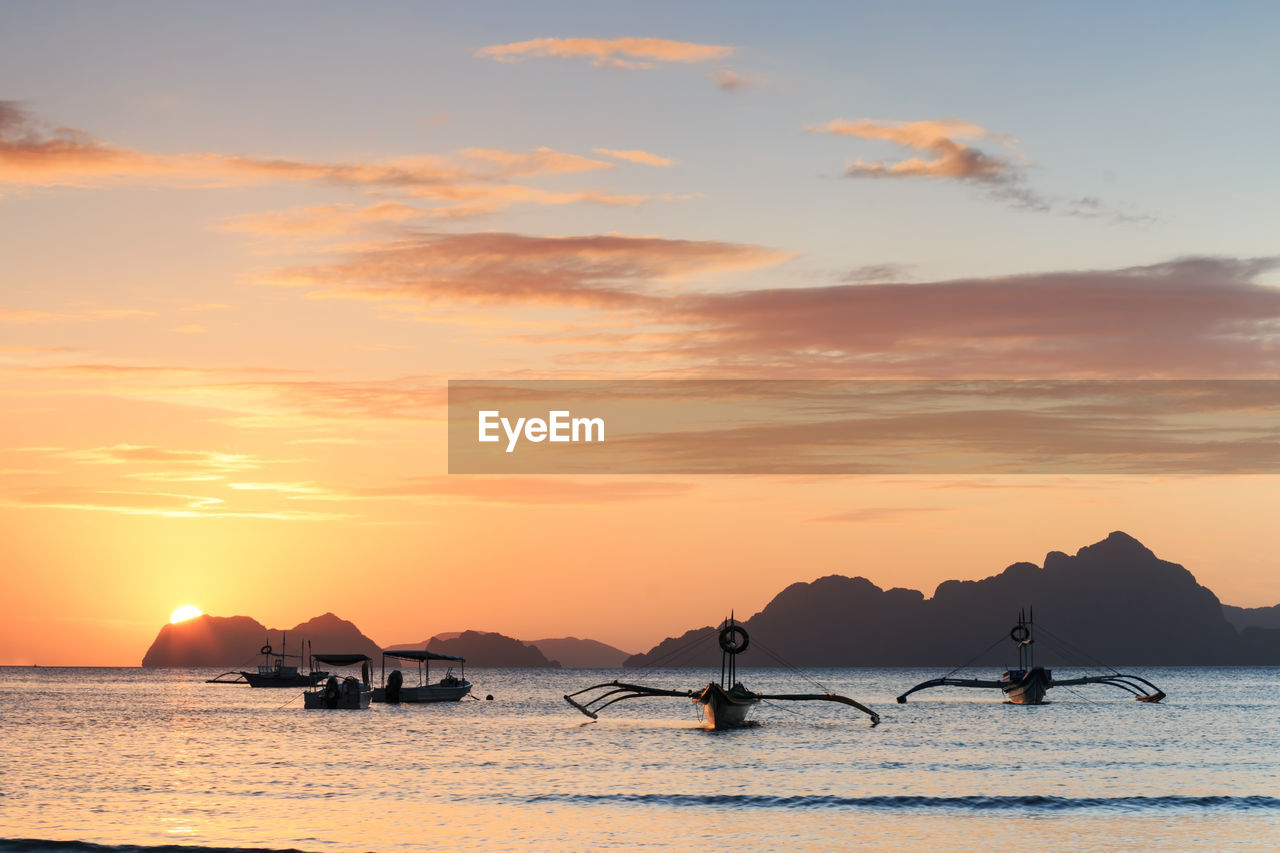 Boats in sea by mountains against cloudy sky during sunset