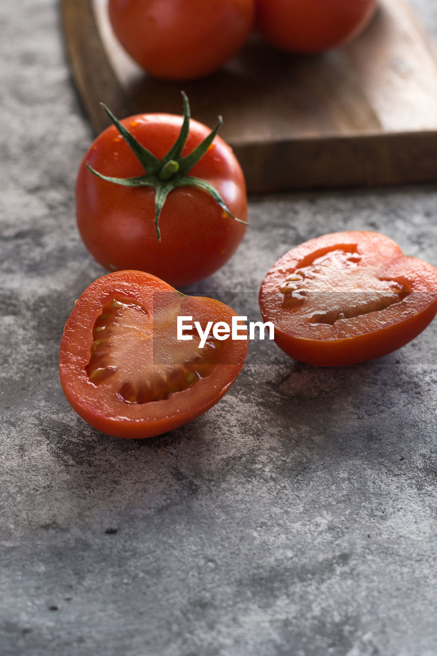Halved and whole fresh tomatoes placed wooden board on rough gray table during food preparation in kitchen