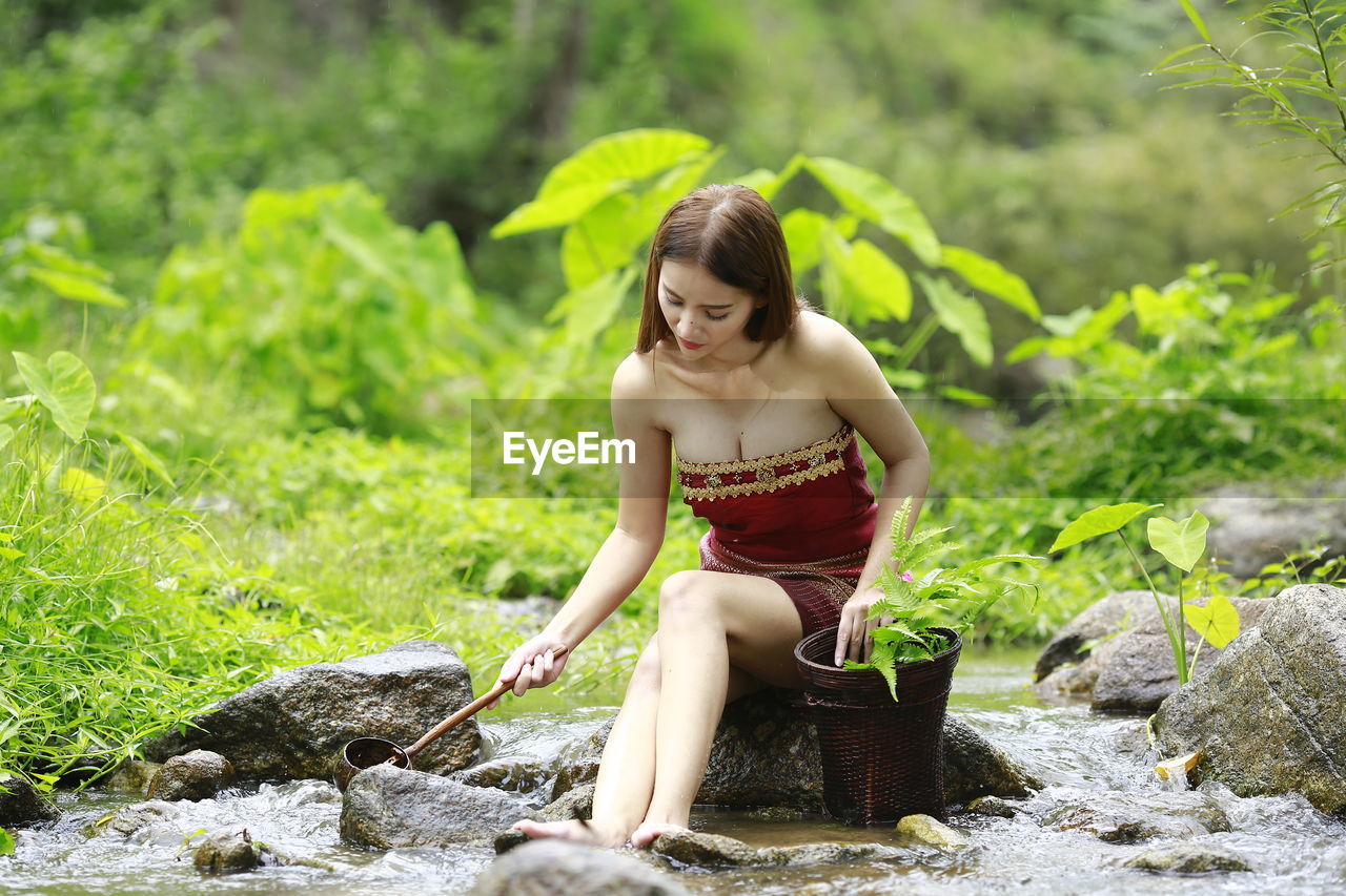 YOUNG WOMAN SITTING ON ROCK BY WATER