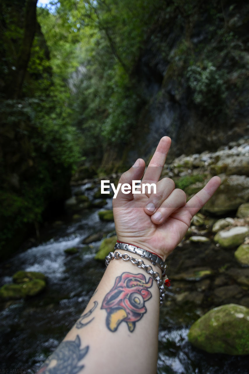 Close-up of tattooed hand gesturing against stream flowing in forest