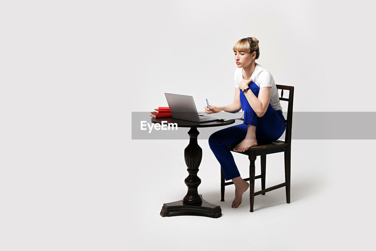 portrait of smiling young woman using laptop while sitting on chair against white background