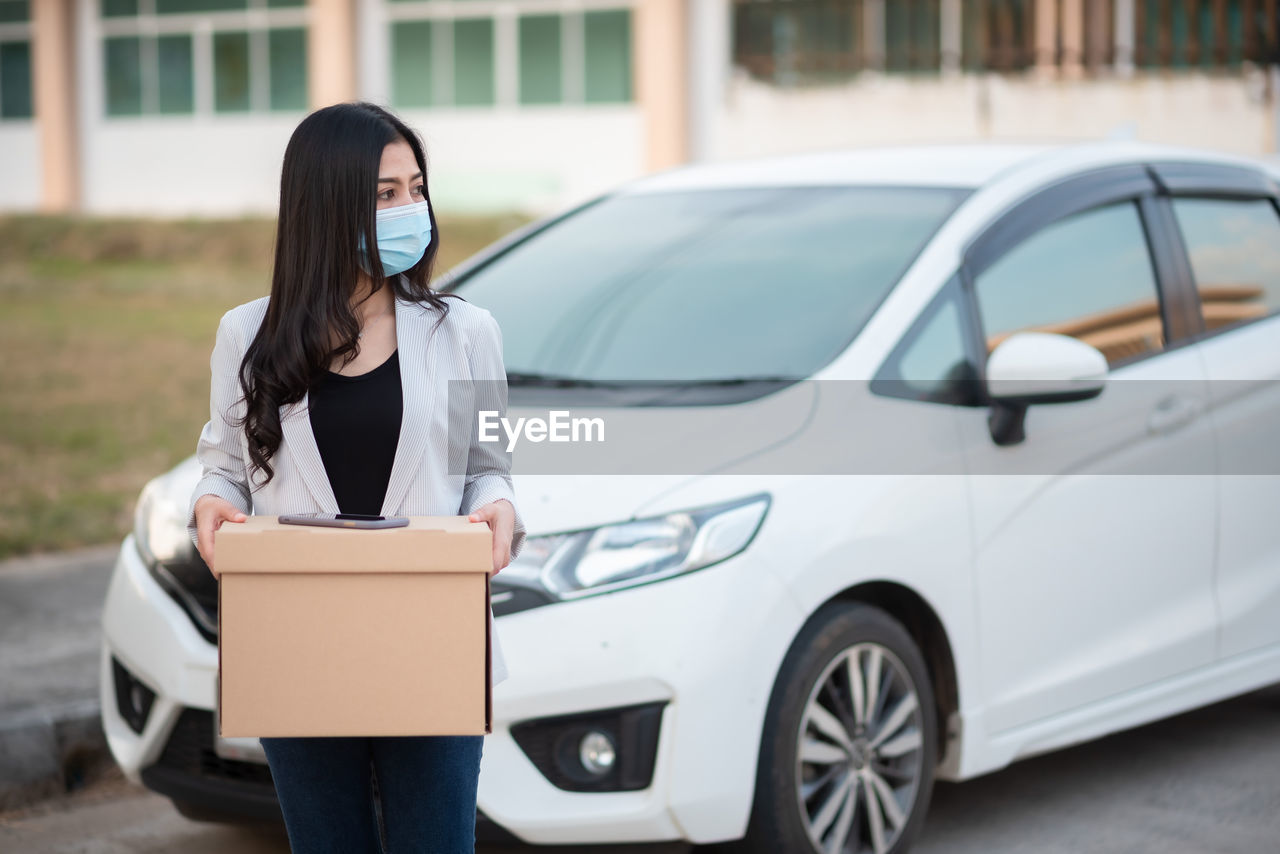 Woman wearing mask holding cardboard box standing by car