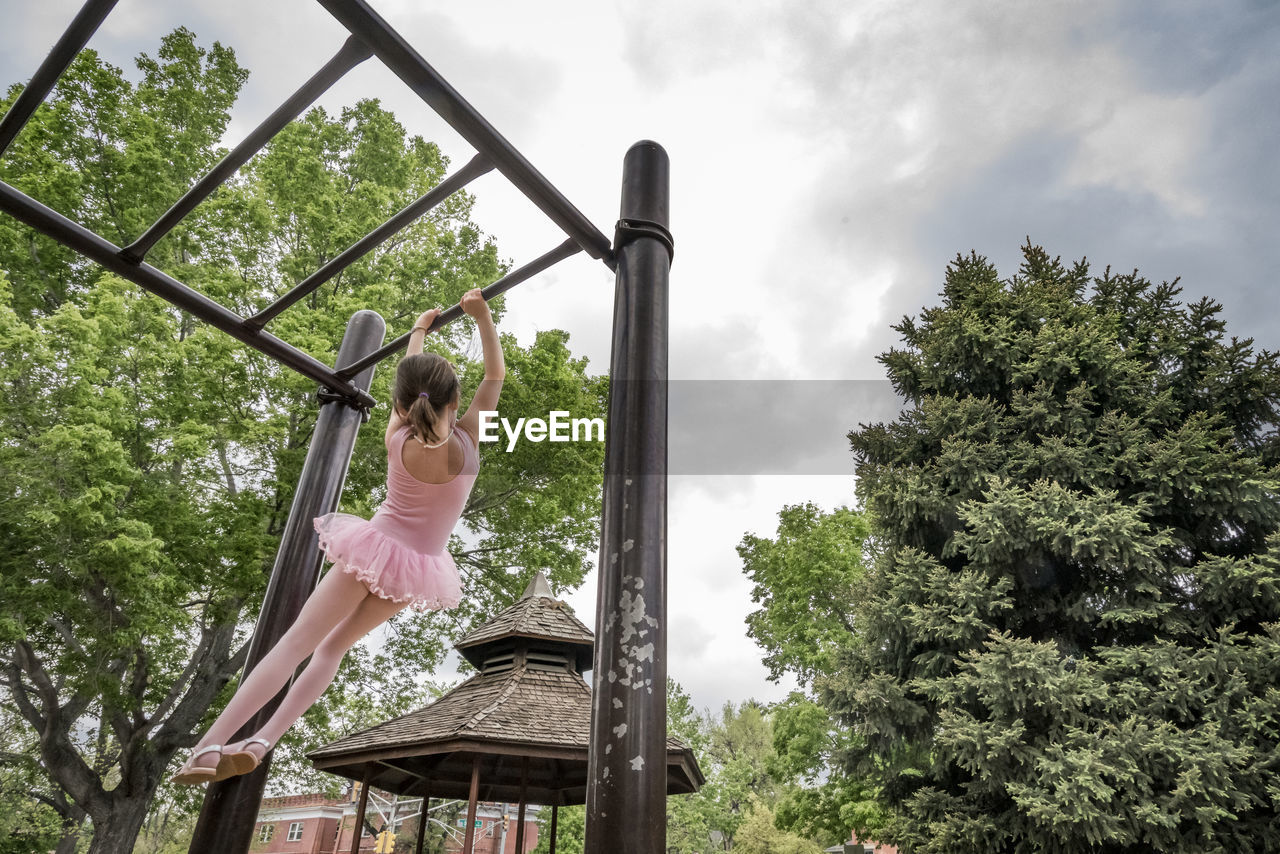 Rear view of girl in ballet costume hanging on monkey bars against cloudy sky at playground