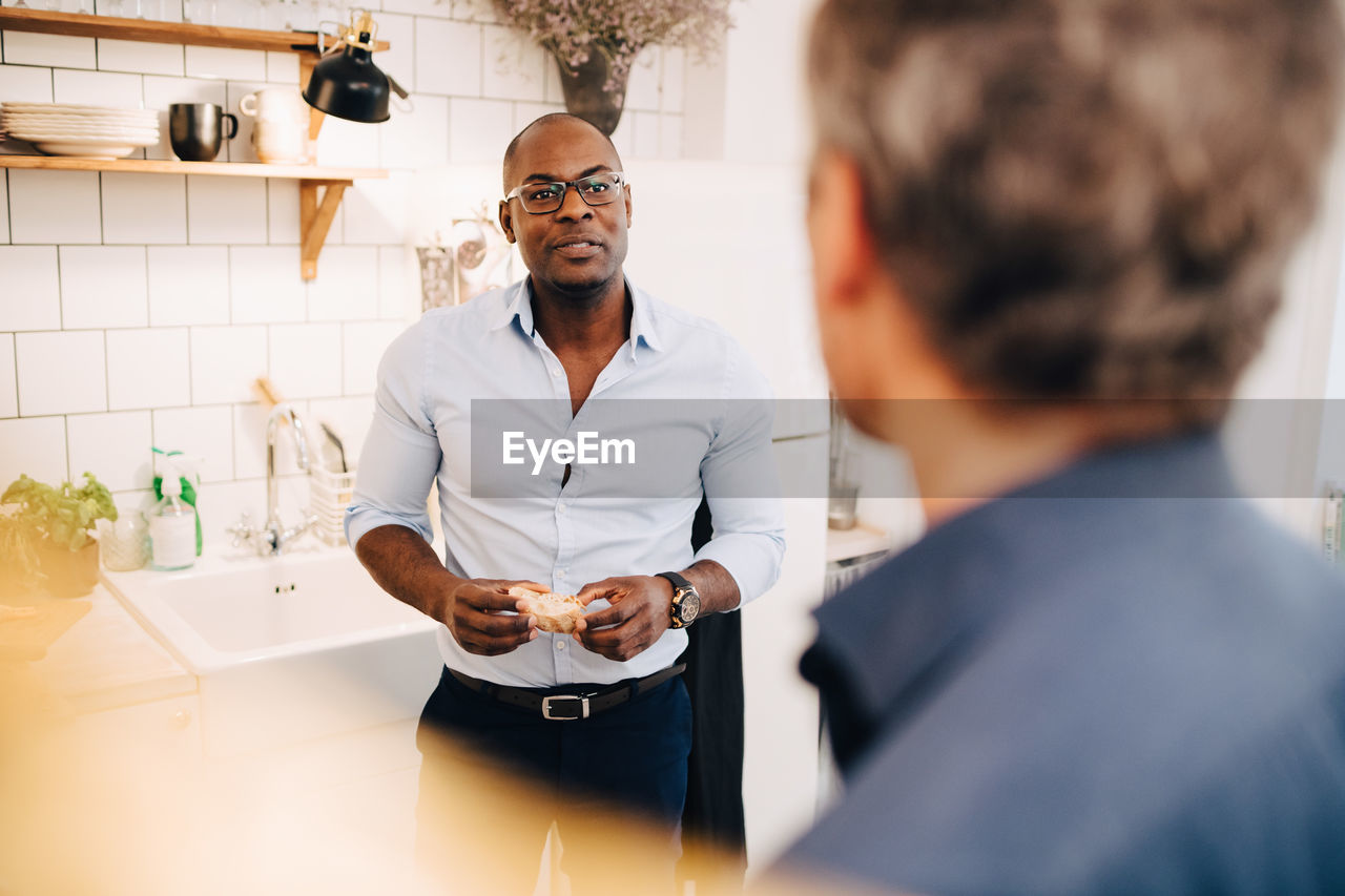 Man holding bread slice while talking to friend in kitchen at home